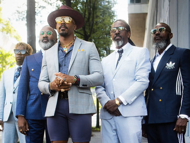 Five men standing together on a city sidewalk wearing stylish suits and sunglasses. The center man wears a light gray blazer, dark shorts, and brown loafers, while the others wear blue, white, and navy suits.